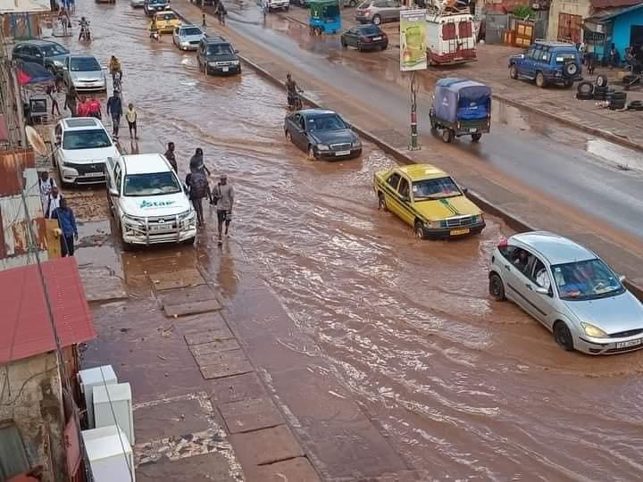 überflutete Straße in Gambia während Regenzeit, mit Autos und watenden Menschen