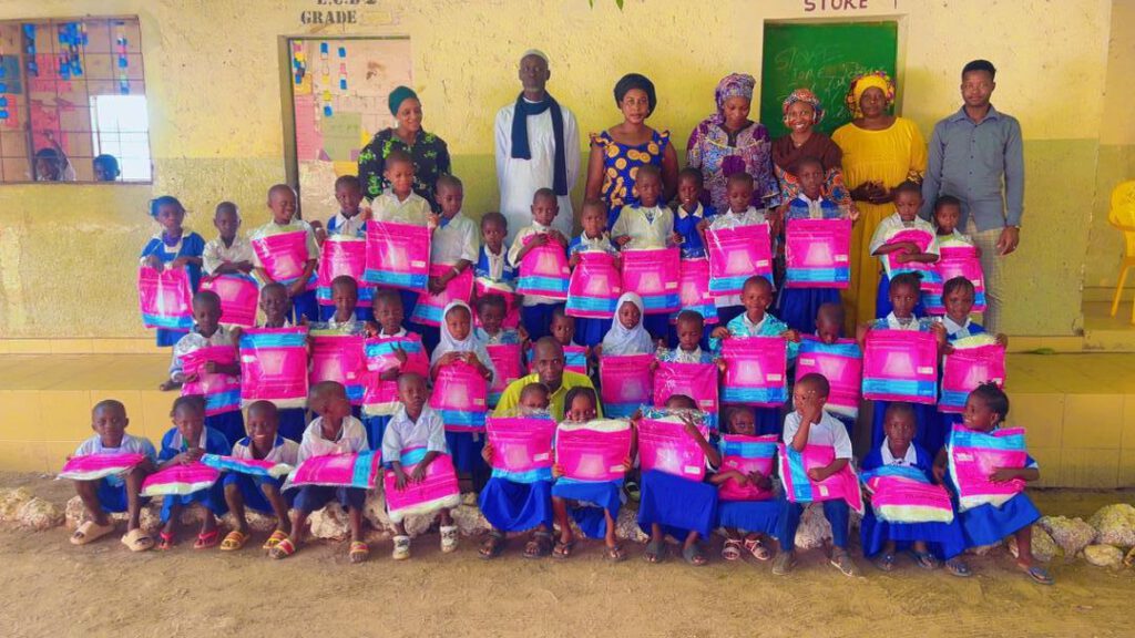 Drei Reihen sitzende Kinder mit Moskitonetzen auf dem Schoß, dahinter eine Reihe Lehrpersonal, vor Schulgebäude Medina